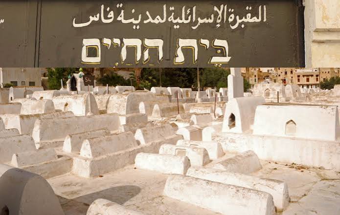 The entrance to the Jewish cemetery of Fes — the sign above reads in Arabic 'Al-Maqbara al-Isra'iliyya li-Madinat Fas' (Jewish Cemetery of the City of Fes) and in Hebrew 'Beit HaChaim' (House of the Living), the traditional Jewish name for a cemetery. White plaster tombs extend behind the entrance in dense rows.